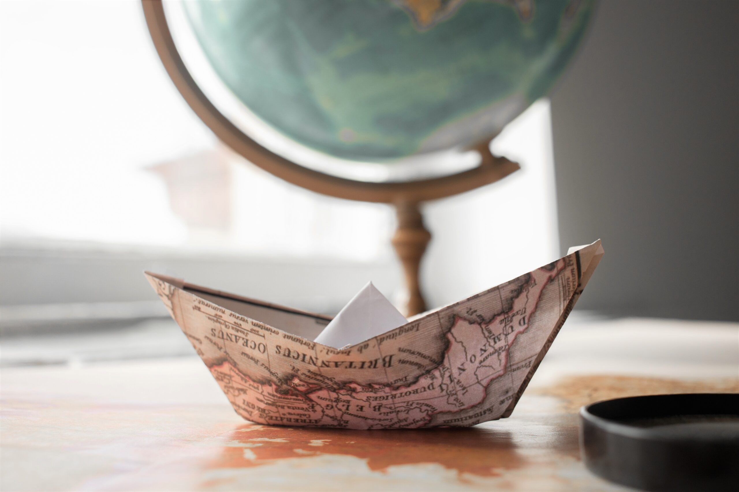 A paper boat made from a map is resting on a wooden table with a globe in the background.