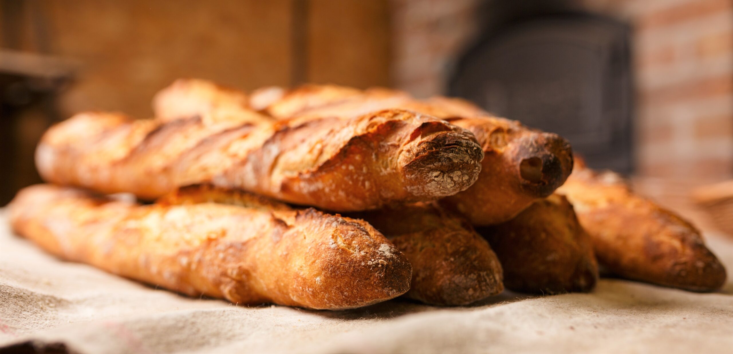 Five freshly bakes baguettes are sitting on a wooden table covered with a thick white linen tablecloth.