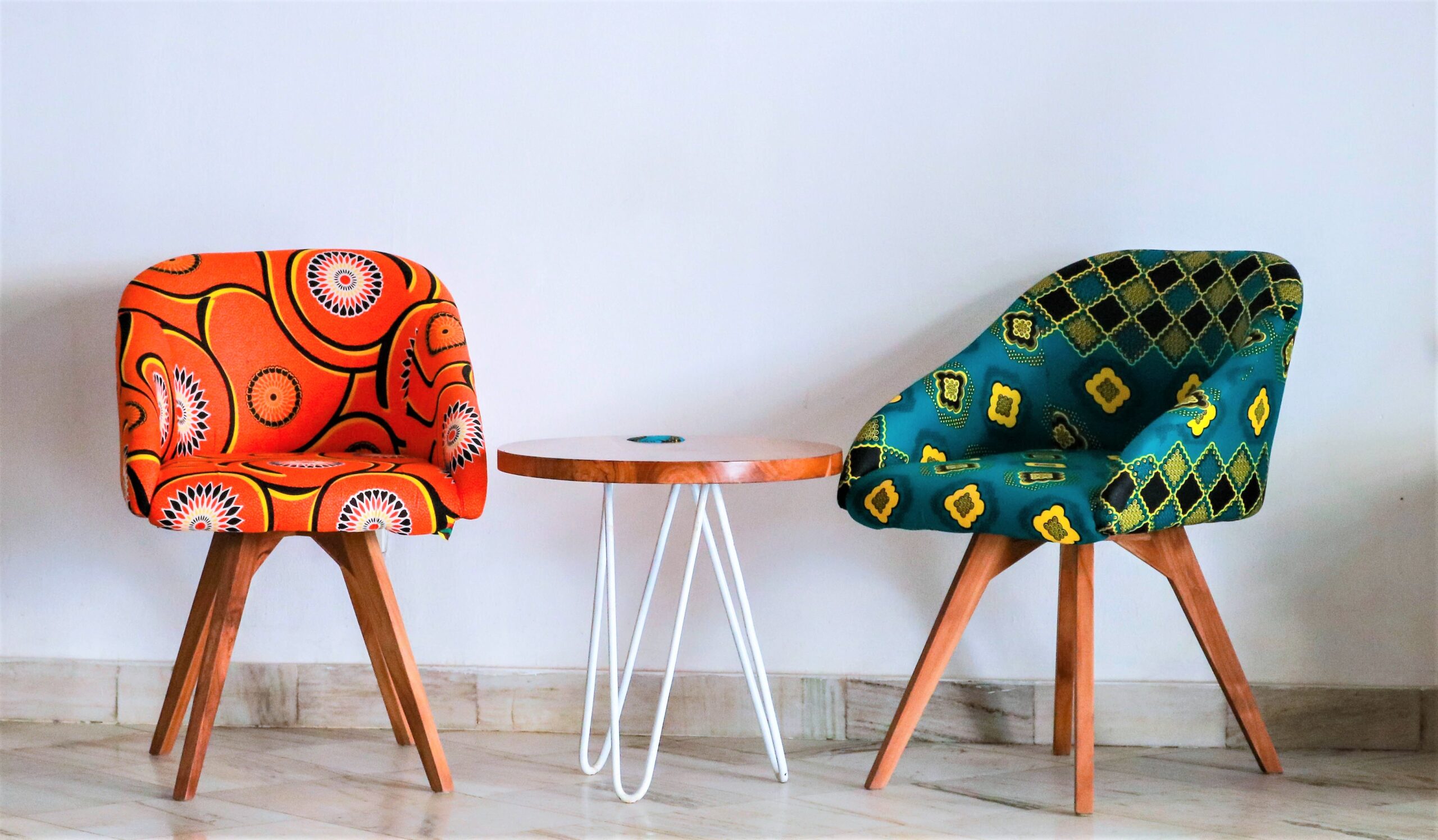 Two brightly reupholstered second hand chairs are set in front of a white wall with a small hairpin legs coffee table in between.