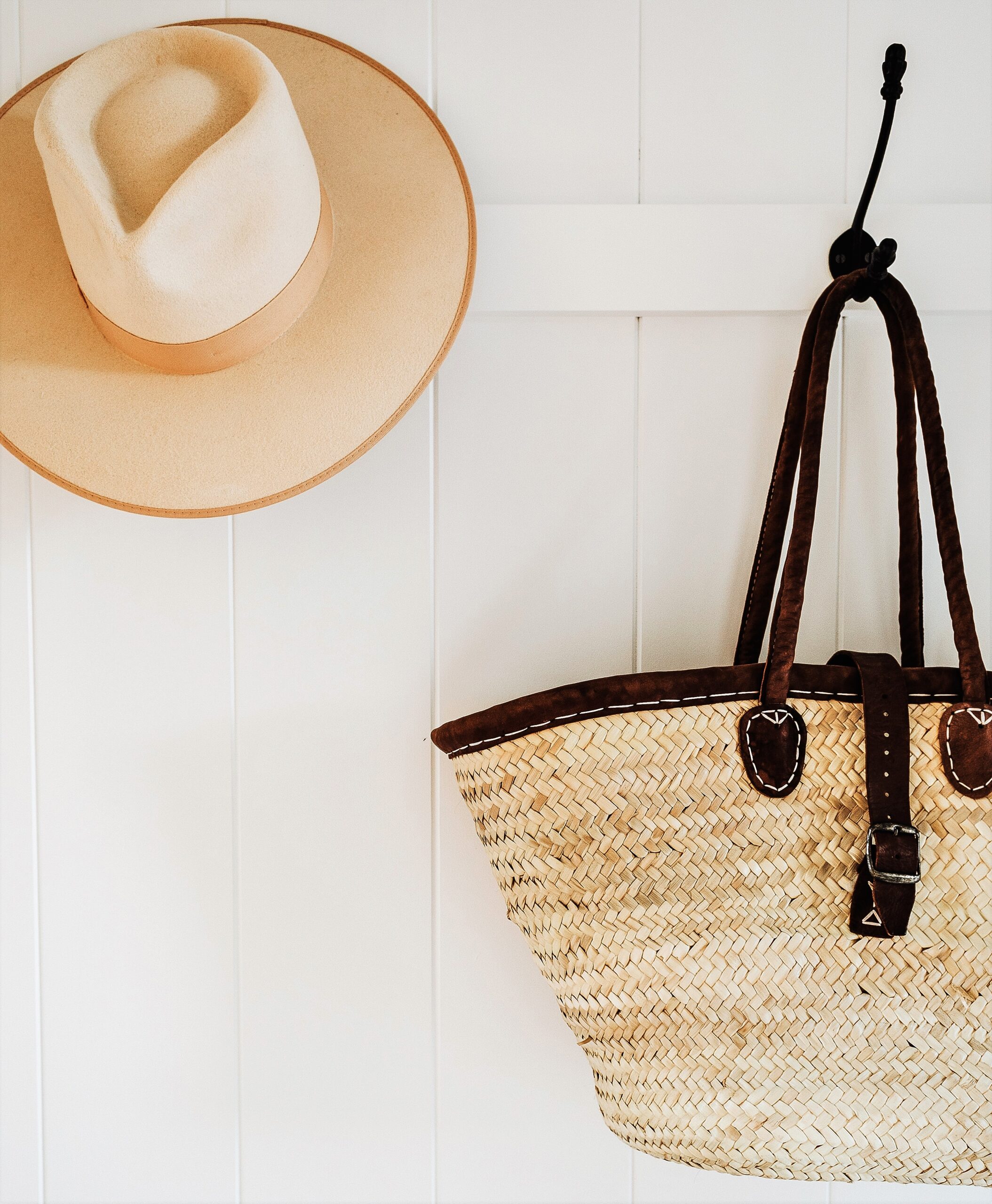 Two black iron hooks are set on a white wall, holding a weaved basket and a wide-brimmed Australian outback hat in light color.