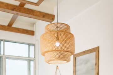 A stunning wicker pendant light is brightening a white bedroom with visible clear wood beams on the ceiling and light pouring in from an unadorned window.