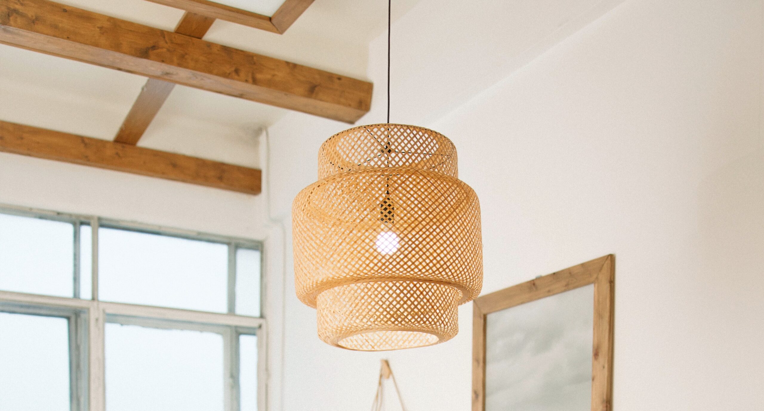 A stunning wicker pendant light is brightening a white bedroom with visible clear wood beams on the ceiling and light pouring in from an unadorned window.