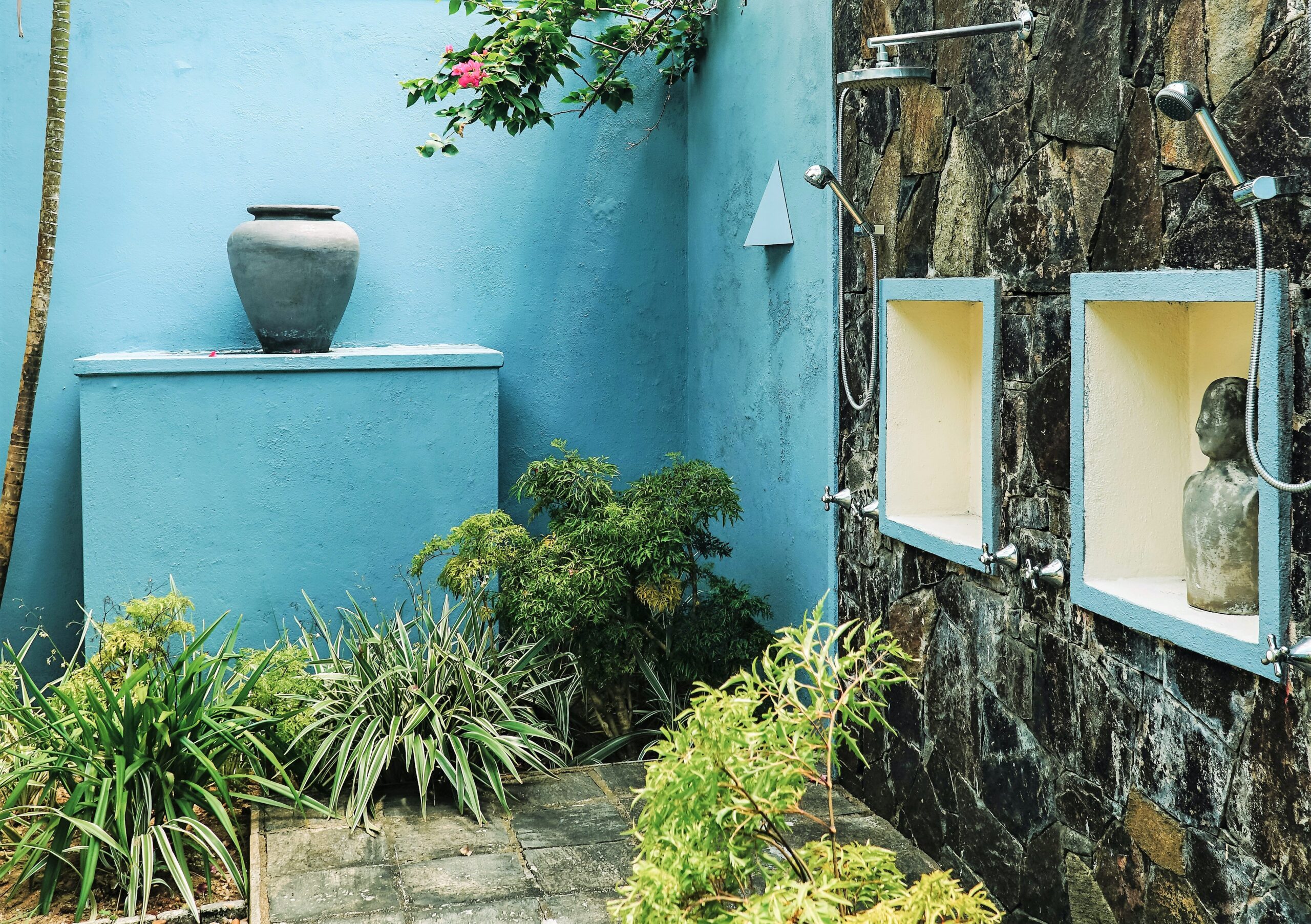 An outdoor shower setting shows bright blue concrete walls and a well drained mix of gravel and stone ground with lots of lush green plants around.