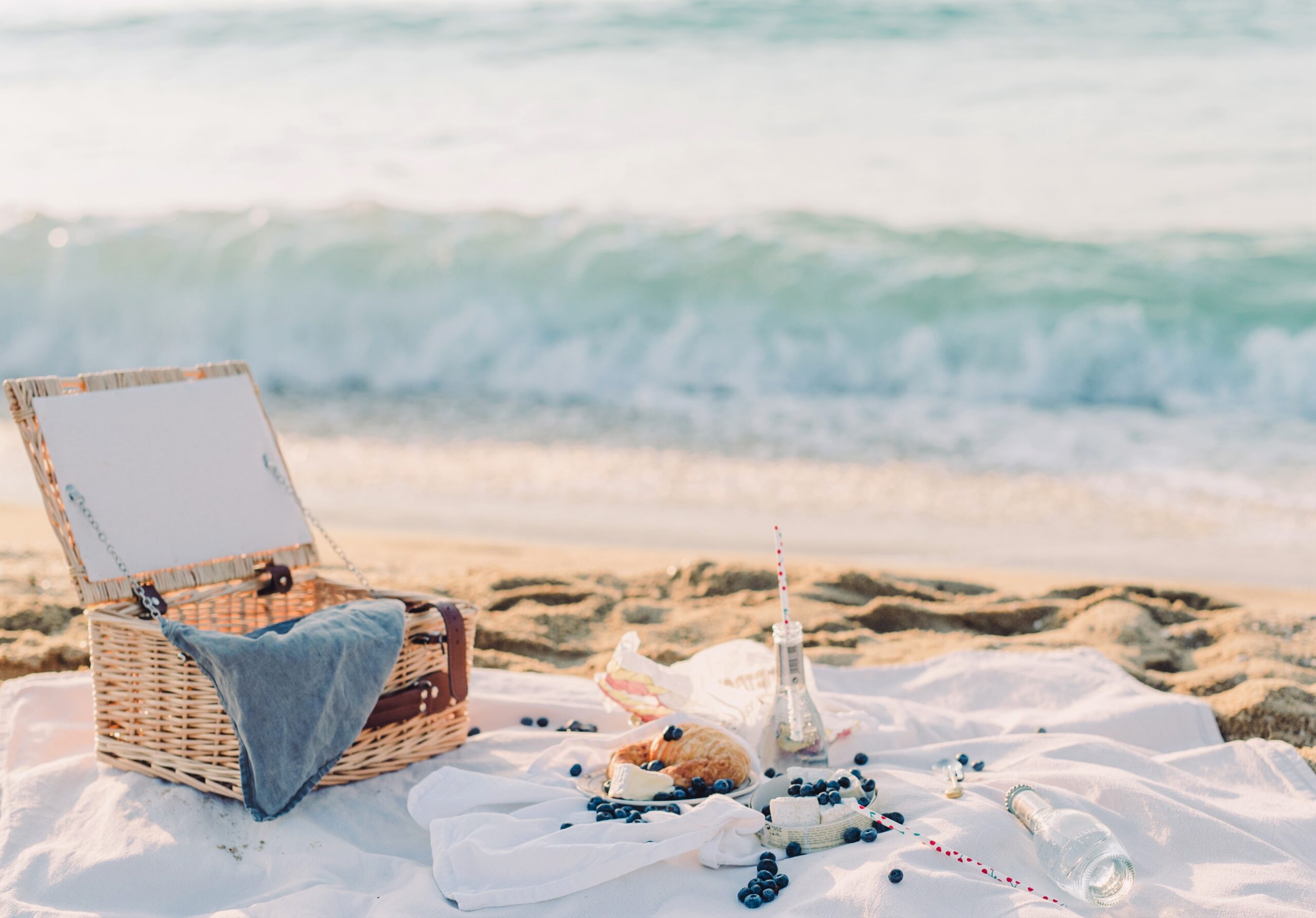 A picnic is set on a sunny beach, white cloth on the sand with food.