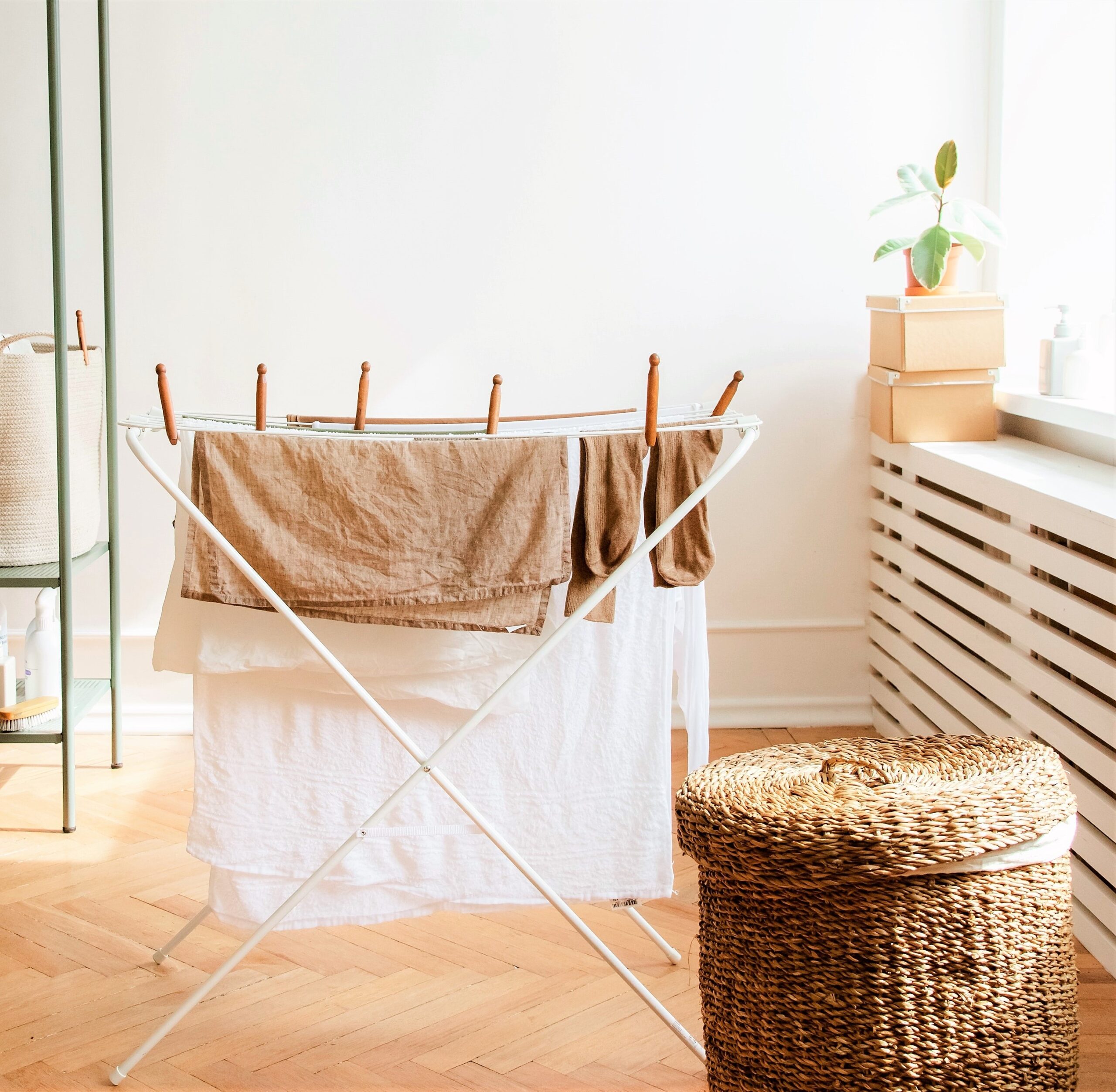 A laundry basket is set on a light wooden floor with a rack of drying clothes in the background.