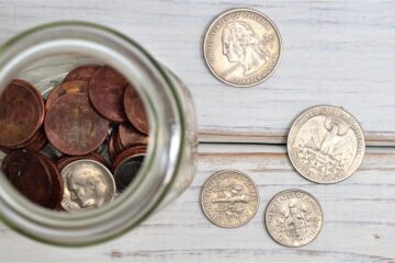 A jar filled with coins is sitting on a whitewashed board table.