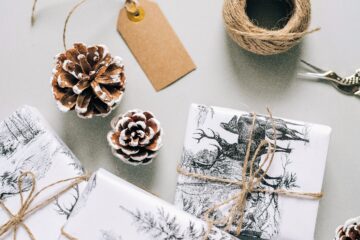 Various presents wrapped in white sturdy paper with bold black prints are set on a table with some pine cones beside, a vintage pair of scissors a ball of twine and cardboard tag.