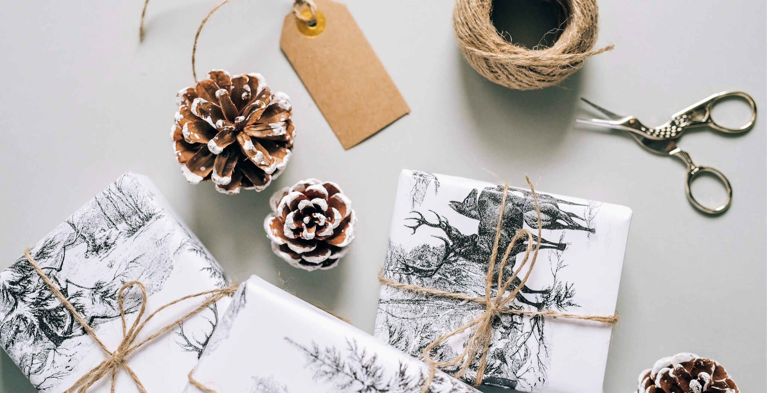 Various presents wrapped in white sturdy paper with bold black prints are set on a table with some pine cones beside, a vintage pair of scissors a ball of twine and cardboard tag.