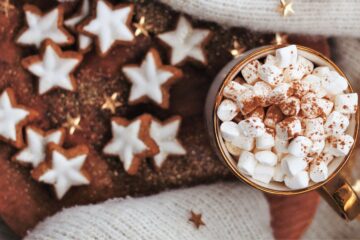 A cup of hot cocoa with lots of mini marshmallows is resting on the sleeve of a white sweater, on a couch. Gingerbread cookies in the shape of stars and decorated with white