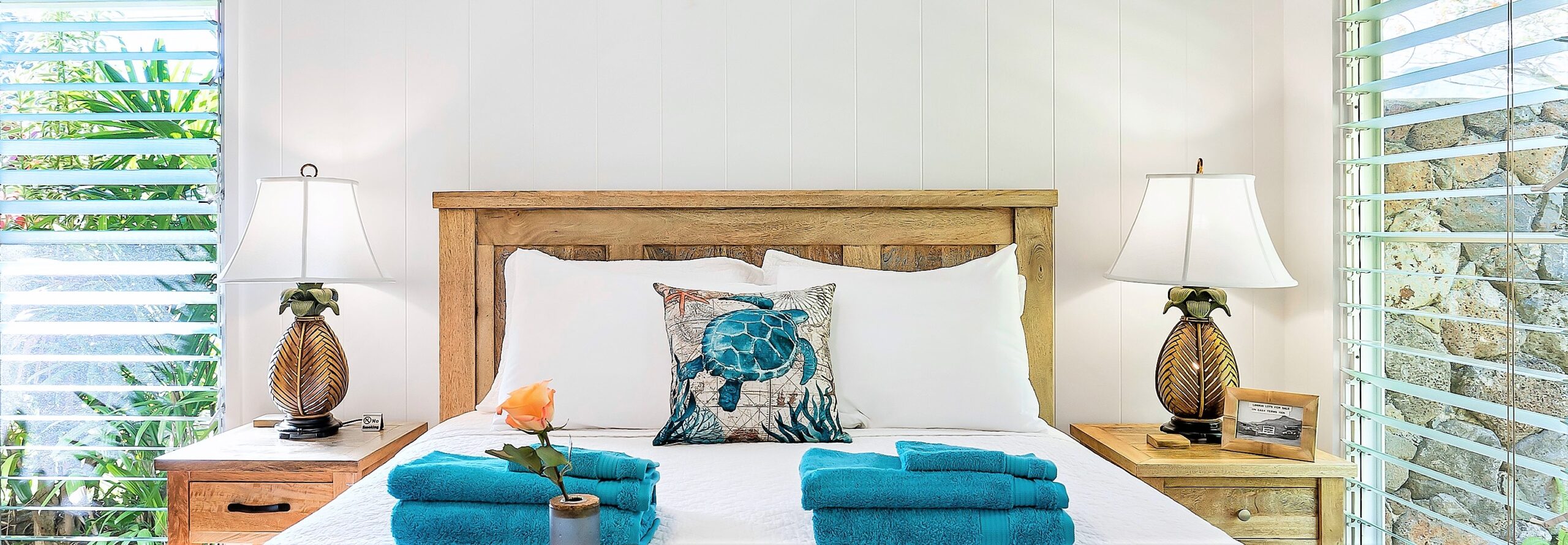 A bed is set on a back white wall with beep turquoise towels prepared on the cover and a cheerful pillow depicting a turtle. Two long windows on each side bright lots of saturated light into the room.