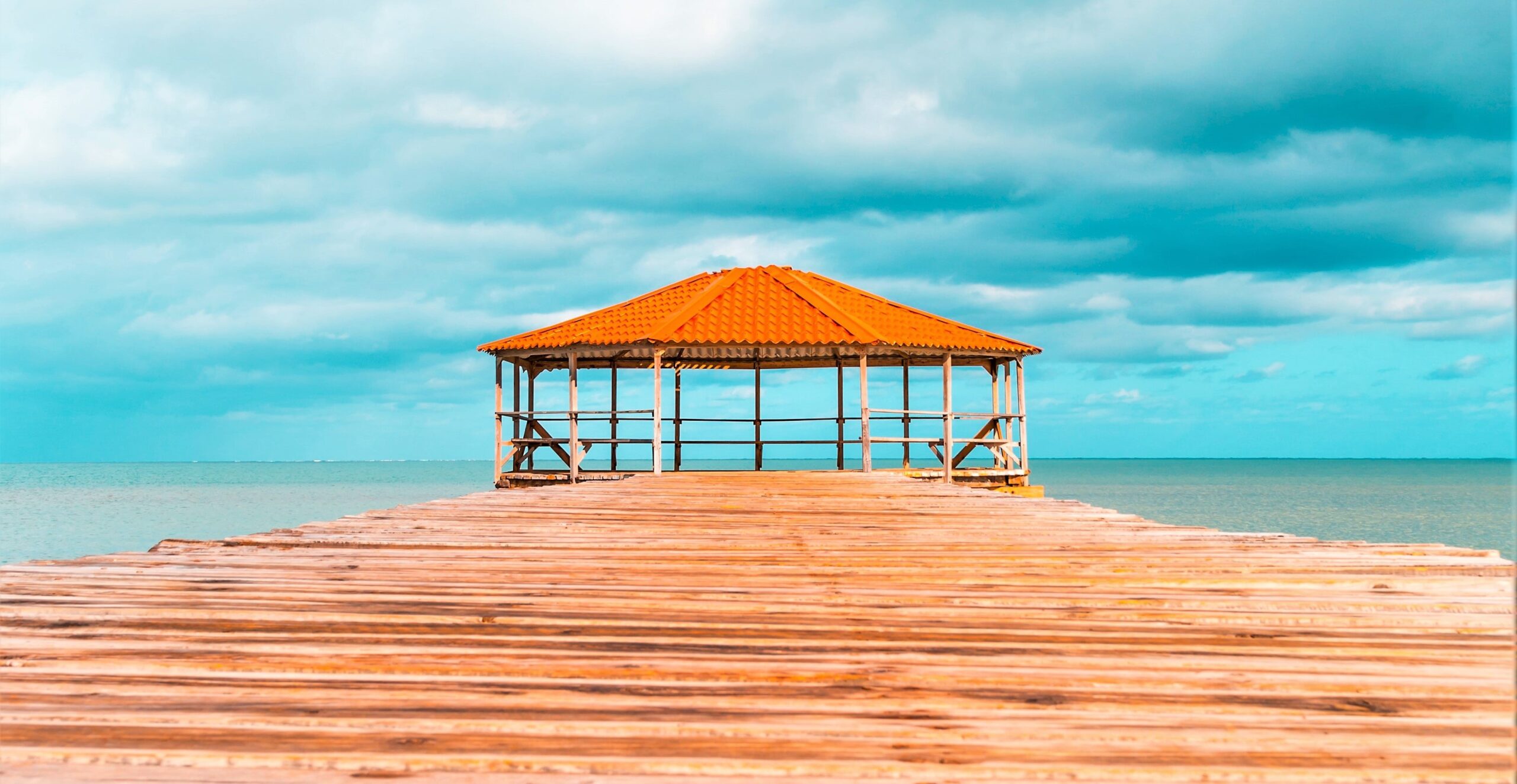 A pergola is set at the end of a boardwalk by the ocean, with a deep, saturated orange roof. The sky is bright turquoise and the boardwalk is made of natural rich toned wood planks.