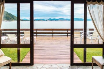 A set of sliding doors is opened on a deck with smooth transition between the inside of the home to the outside for a seamless indoor-outdoor living and with a bright coastal view in the background.