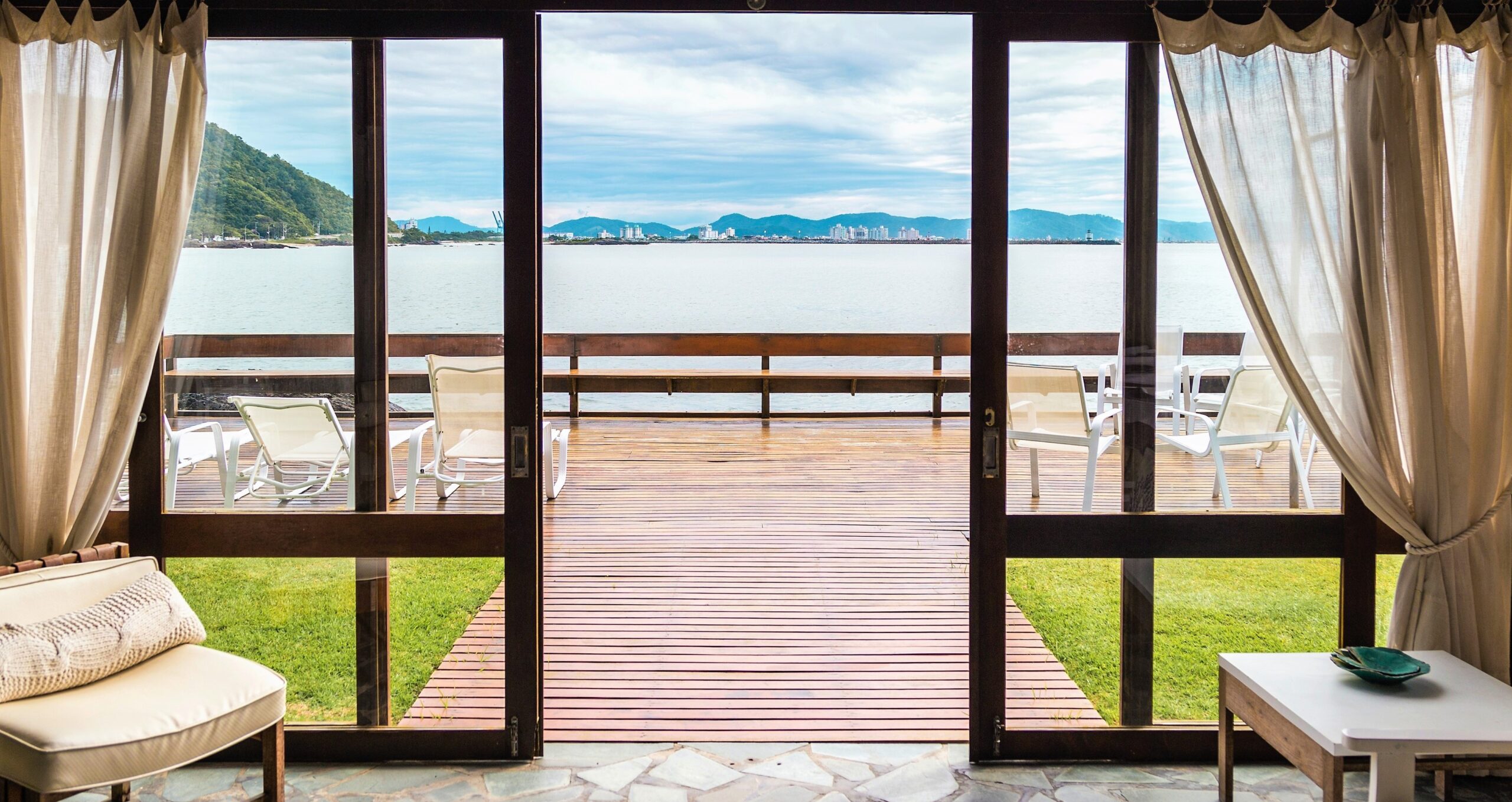 A set of sliding doors is opened on a deck with smooth transition between the inside of the home to the outside for a seamless indoor-outdoor living and with a bright coastal view in the background.