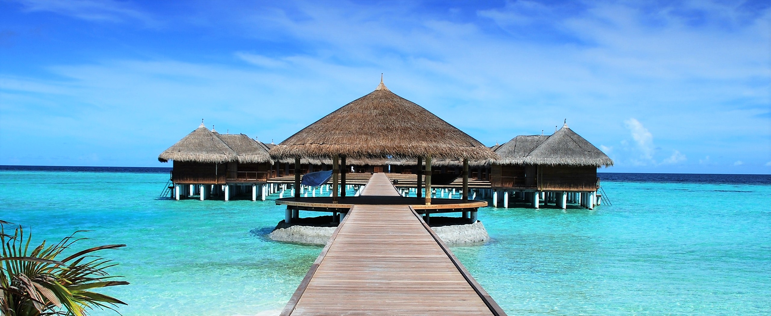 A wide and sturdy wood boardwalk leads from a soft sandy beach strait to a series of circular and open bungalows set on stilts right in the bright turquoise ocean.