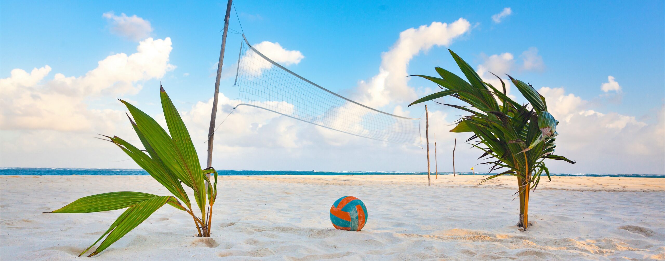 A volleyball is set under a net on a white sand beach on a sunny day.
