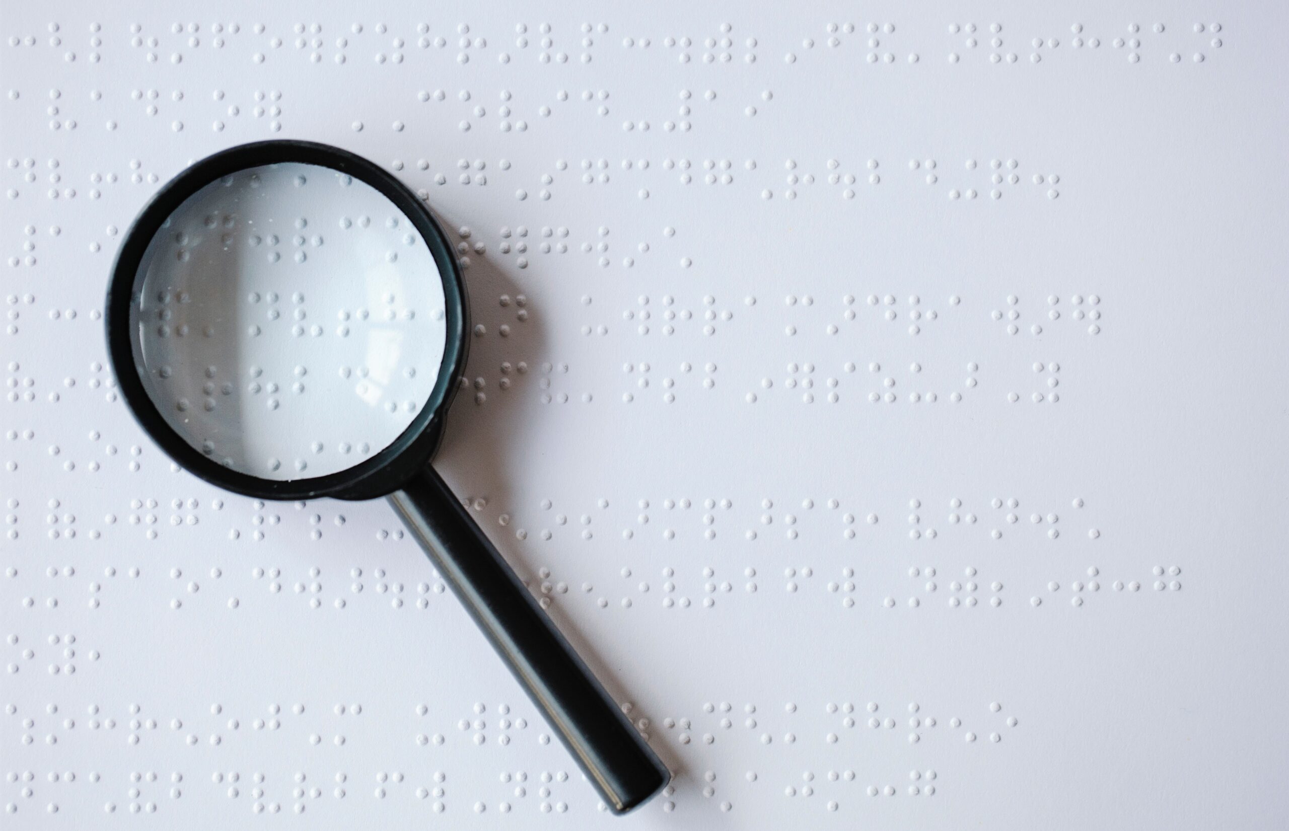A white paper sheet with Braille inscriptions is showing through a simple back magnifying glass.